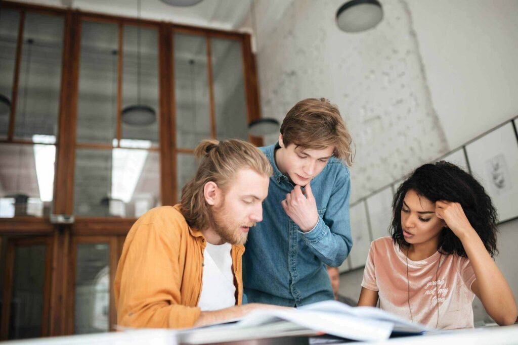 Tres estudiantes sentados en una mesa de madera revisando apuntes y libros juntos.
