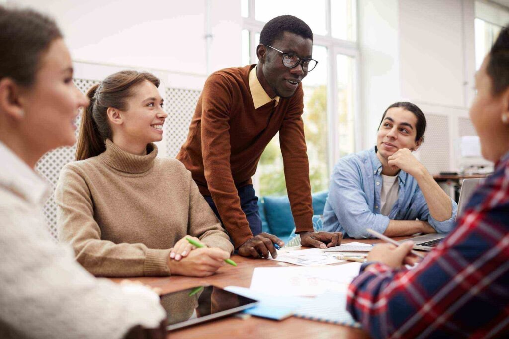 Grupo de cuatro jóvenes sentados alrededor de una mesa, escribiendo en cuadernos y conversando en un ambiente de aula.