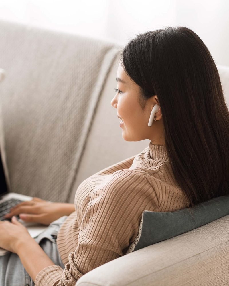 Mujer joven usando una laptop en un sillón, con una libreta y bolígrafo a su lado.
