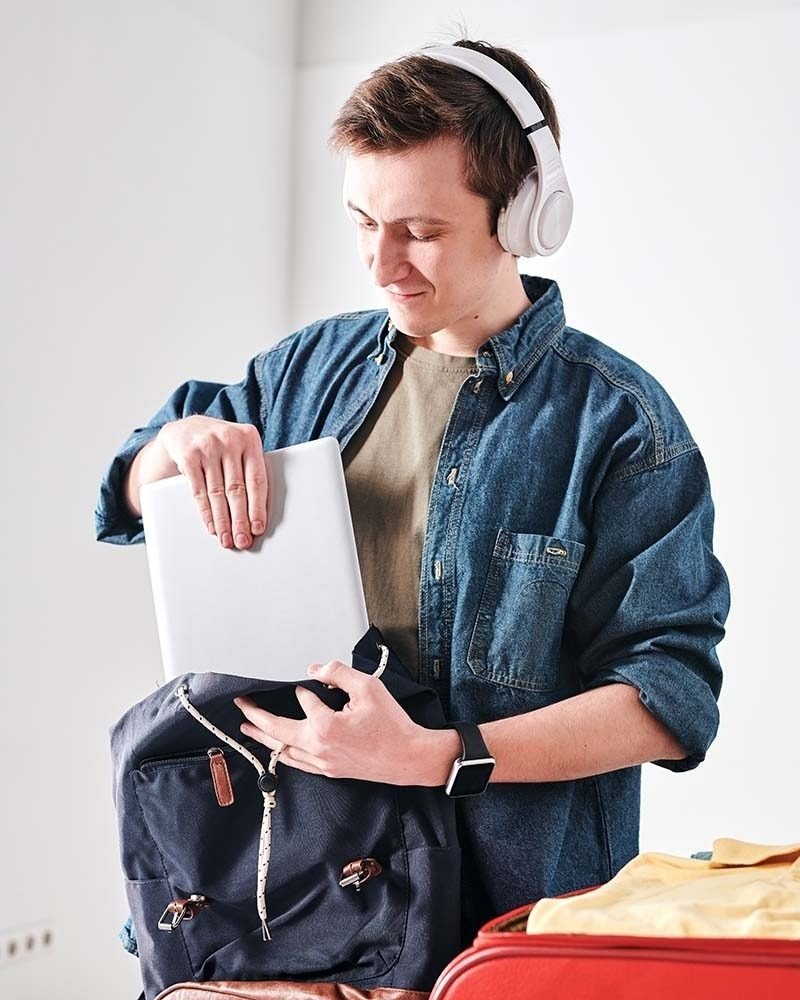 Joven con auriculares guardando libros y cuadernos en una mochila, junto a una laptop.