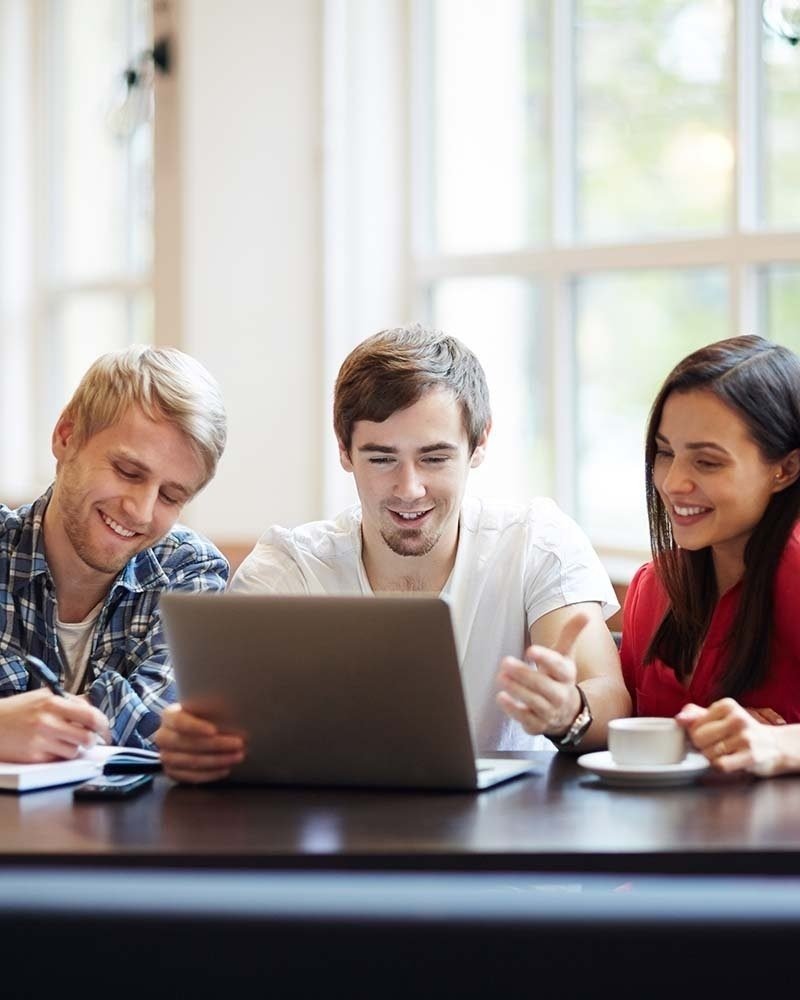 Dos jóvenes y una joven mirando juntos una laptop y sonriendo mientras estudian.
